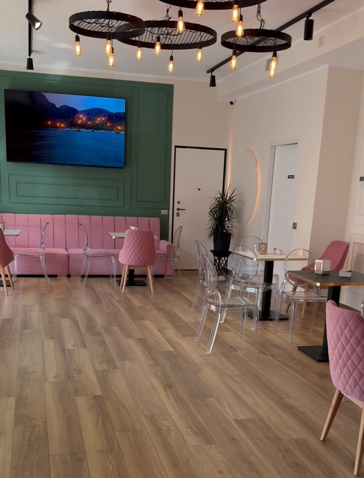 Modern lounge interior with pink velvet seating, green accent wall, mounted TV, clear chairs, wooden flooring, and industrial pendant lighting