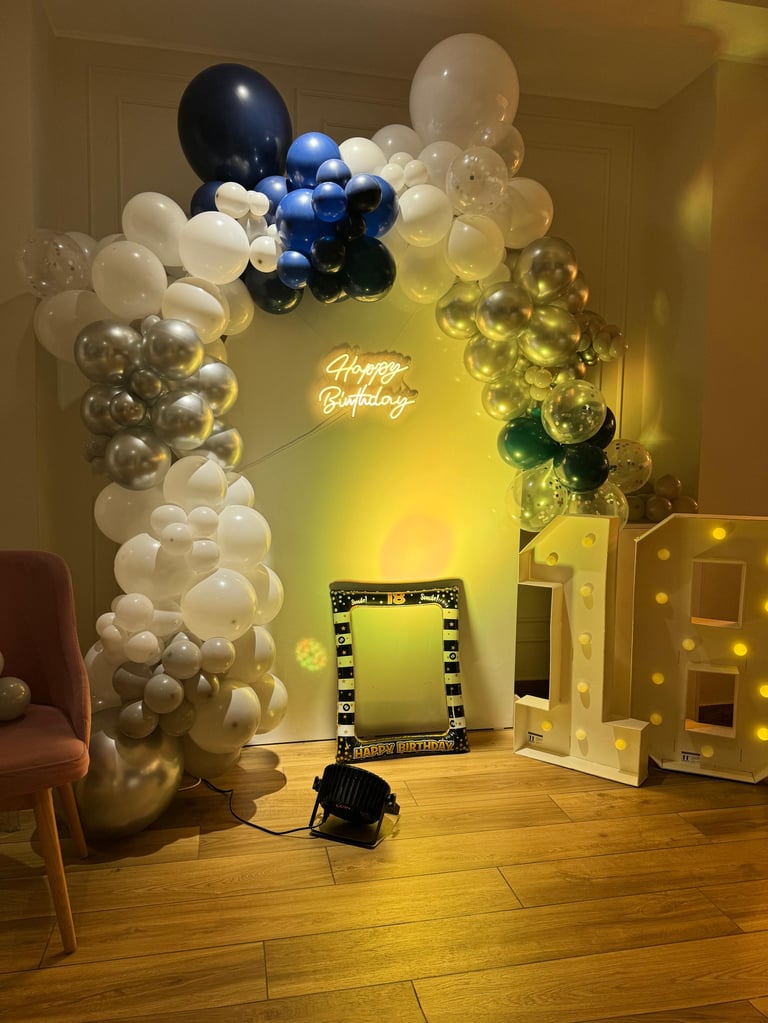 Birthday party setup with balloon garland arch in blue, white, and gold tones, illuminated Happy Birthday neon sign, and vanity mirror with lights