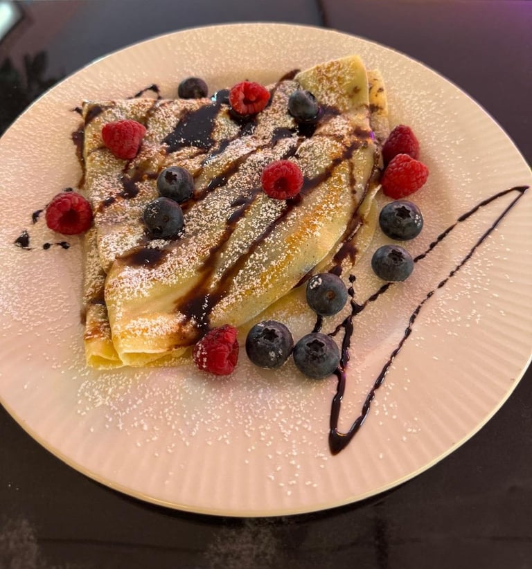 Crepes dusted with powdered sugar, topped with fresh raspberries and blueberries, chocolate drizzle, on a white plate