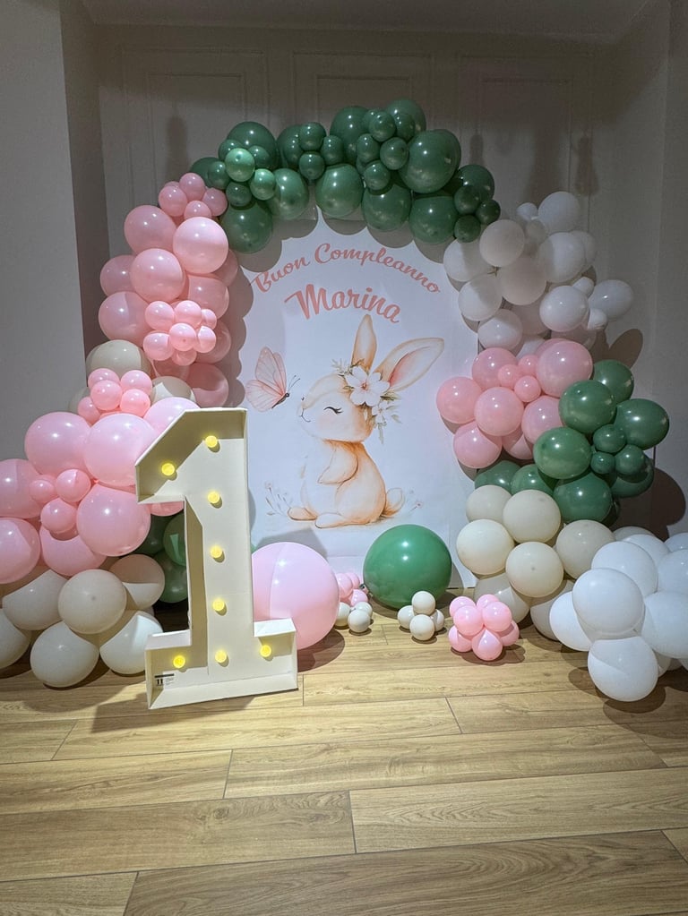 Colorful balloon arch with pink, white, and green balloons surrounding a banner featuring a bunny design and glowing marquee letter, creating a festive birthday setup