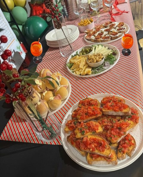Festive party spread with red and white striped tablecloth featuring bruschetta, chips, pretzels, pizza, sandwiches, orange drinks, and green balloons