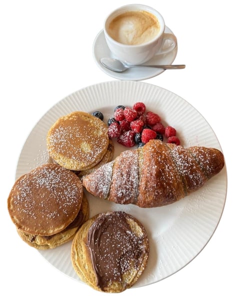 Breakfast plate with powdered sugar-dusted pancakes, croissant, and chocolate pastry, fresh berries, and cappuccino coffee