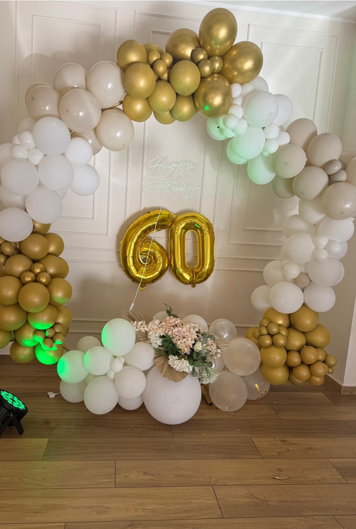 Elegant 60th birthday party decoration with gold and white balloons arranged in an arch, large gold foil number 60, and floral centerpiece on wooden floor